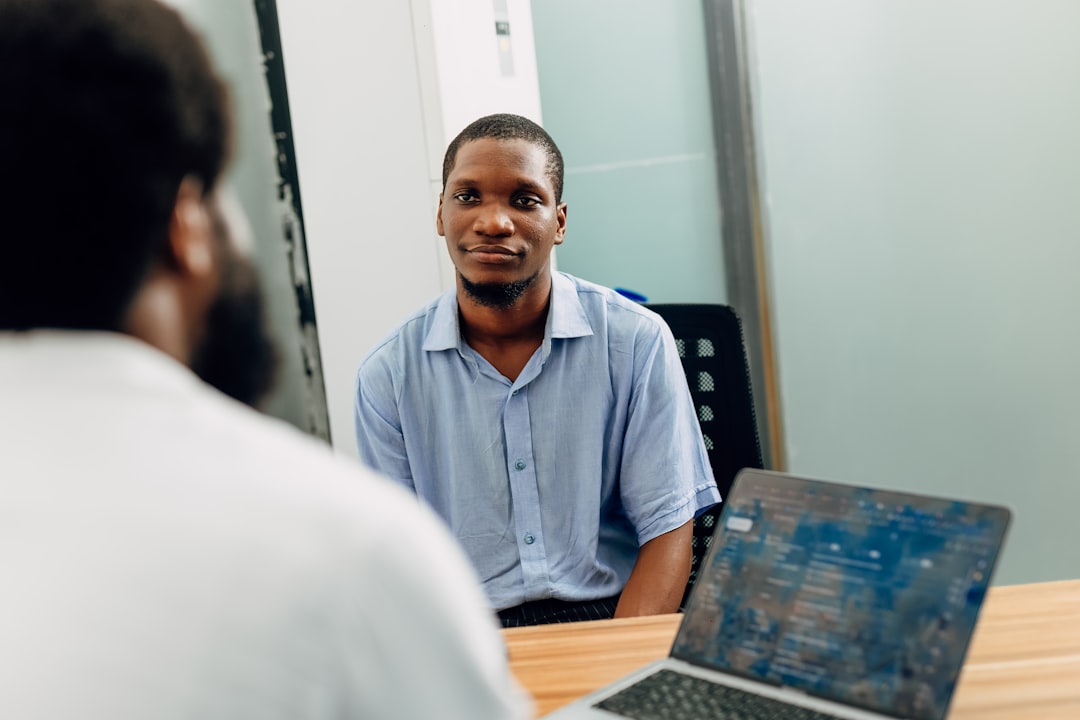 A man wearing a light blue button-down shirt sits in an office at a desk and is listening to the man in front of him. Find more afro-centric images at www.ninthgrid.com